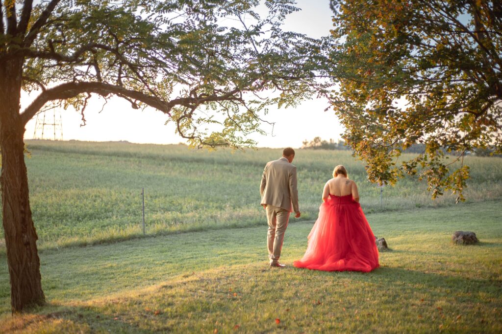 Scène bucolique avec les mariés marchant à travers un champ, la robe rouge de la mariée contrastant avec la verdure environnante.