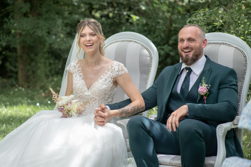 Le couple de mariés riant ensemble, assis sur des fauteuils élégants dans un cadre verdoyant.